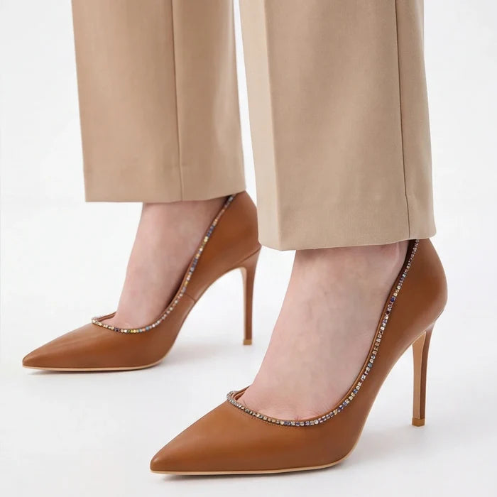 A pair of pointed-toe stiletto pumps in tan leather with a delicate line of small rhinestones along the inner edge, shown on a model wearing beige trousers against a clean white background.