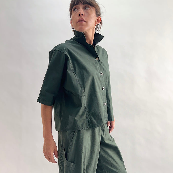 Three-quarter side view of model wearing Eleven Stitch konbu camp shirt looking up, showing stand collar, dark shell buttons, short sleeves, patch pocket, slightly cropped boxy fit, and deep forest green color, styled with matching konbu snap front pant