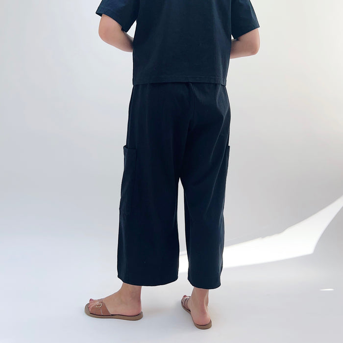 A person standing against a white background, wearing black cotton pasha pants and a black top.