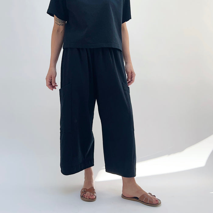 A person standing against a white background, wearing black cotton pasha pants and a black top.