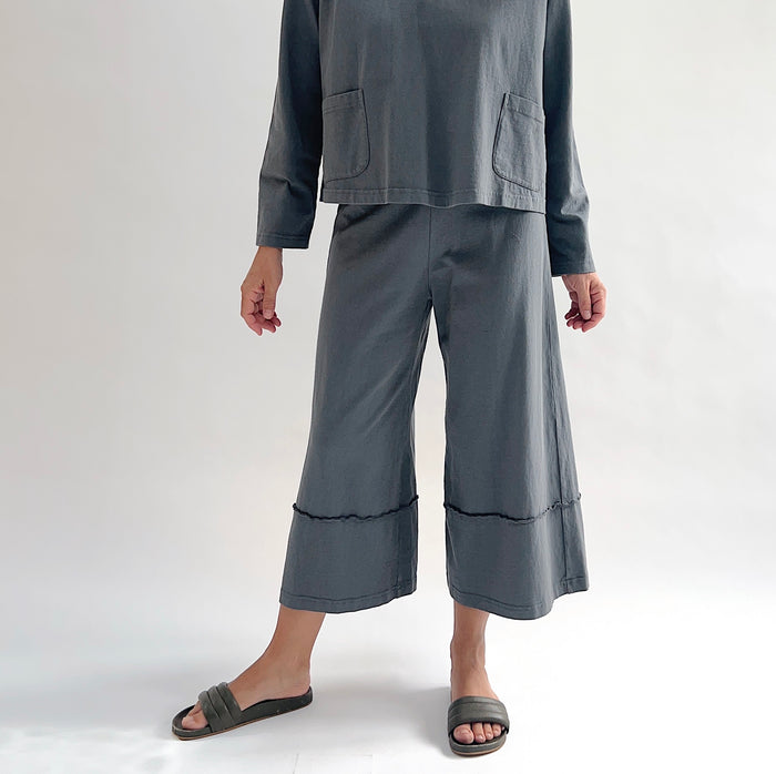 Close front view of model wearing Pacific Cotton dolomite crop full pant, showing elastic waistband, wide flared leg, layered hem panel with exposed seam detail, and washed slate grey color, styled with matching dolomite boxy shirt