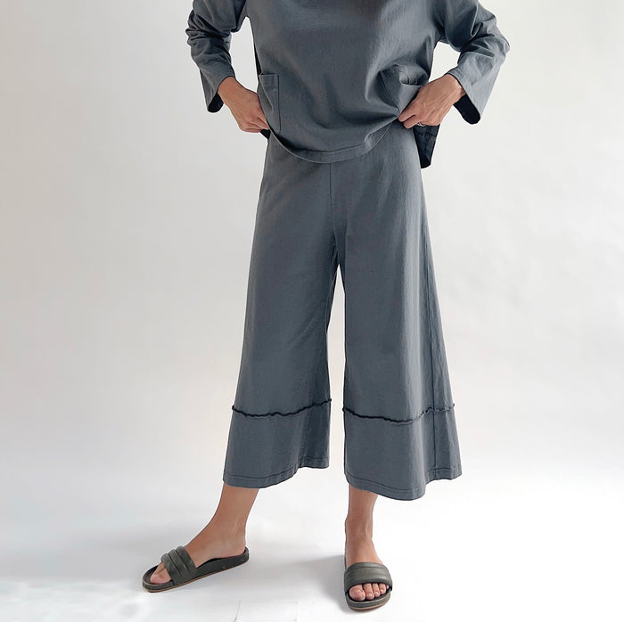 Full front view of model wearing Pacific Cotton dolomite crop full pant with hands on hips, showing elastic waistband, wide flared leg silhouette, layered hem panel, and washed slate grey color, styled with matching dolomite boxy shirt
