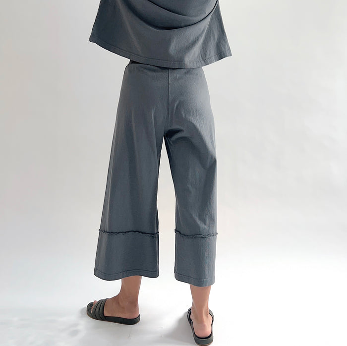 Back view of model wearing Pacific Cotton dolomite crop full pant, showing elastic waistband, wide flared leg, layered hem panel with exposed seam detail, and washed slate grey color, styled with matching dolomite boxy shirt