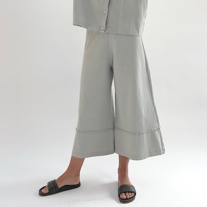 Three-quarter front view of model wearing Pacific Cotton orlaya crop full pant, showing elastic waistband, wide flared leg silhouette, layered hem panel, and cropped length