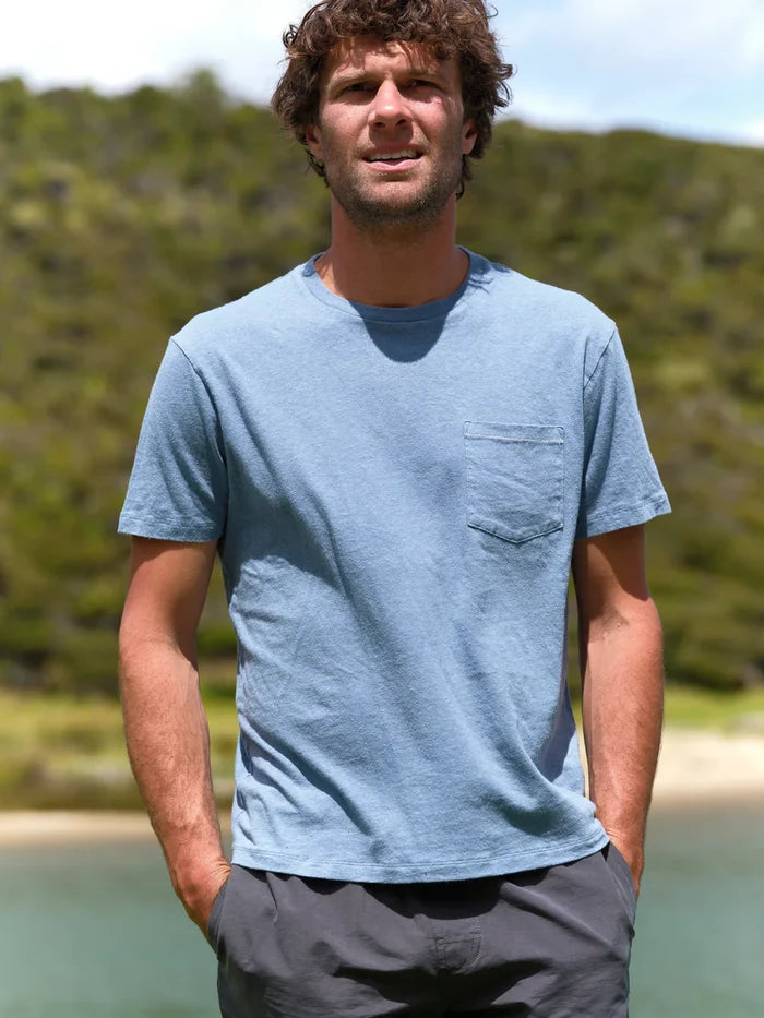 Man wearing a blue t-shirt with a logo standing by a body of water with greenery in the background
