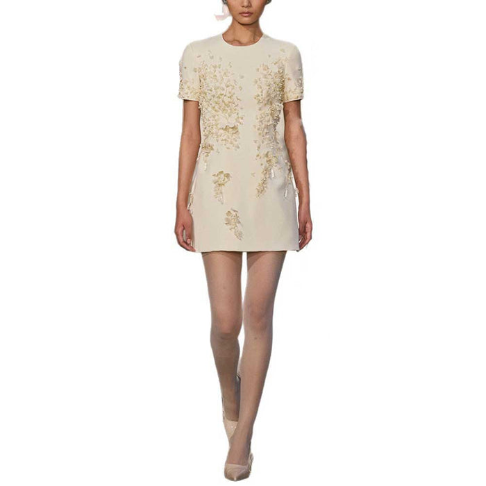 A model wearing a short-sleeve cream-colored mini dress with gold floral embroidery, shown against a plain white background.