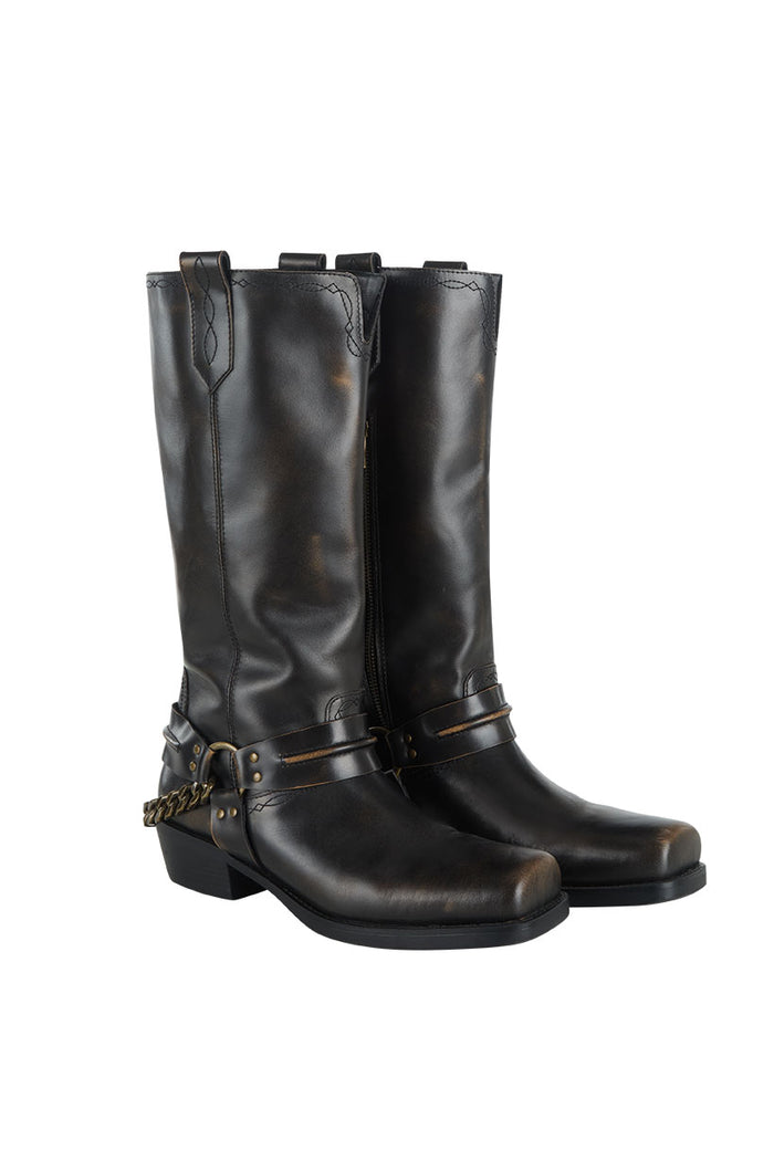 Front flat lay of knee-high vintage black leather biker boots with brass chain harness strap and distressed finish