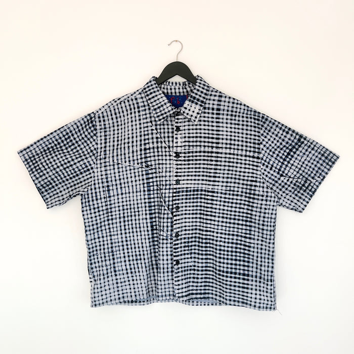 Full front view of the Sudi Shirt in Indigo Grid, hanging on a black hanger against a white wall, showing a point collar, short sleeves, button placket, two chest pockets, and hand-printed indigo and white gingham grid pattern on 100% cotton Batiki fabric