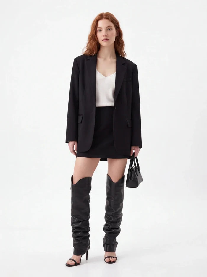 A model stands against a plain white studio background wearing an oversized black blazer, black mini skirt, black thigh-high slouchy boots, a white top, and black heeled sandals, while holding a small black handbag.