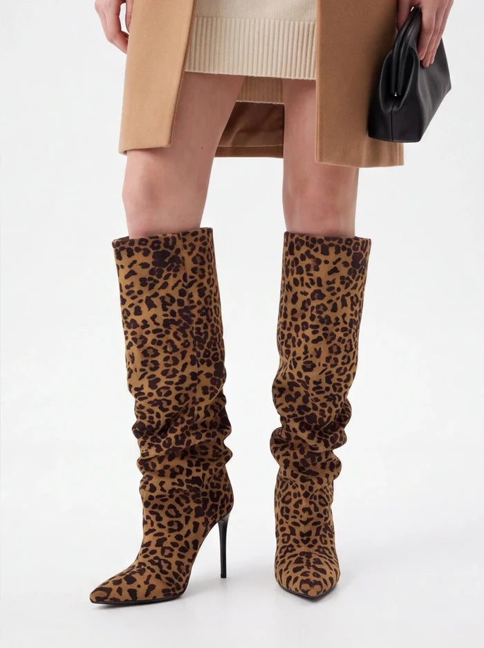 A person wearing knee-high leopard-print heeled boots with a beige ribbed knit top, tan coat, and black handbag against a light gray studio background.
